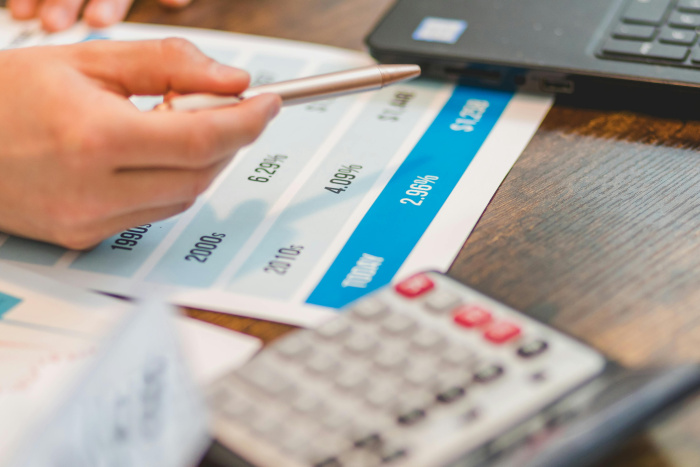 A Hand Holding a Pen over a data sheet calculator and laptop