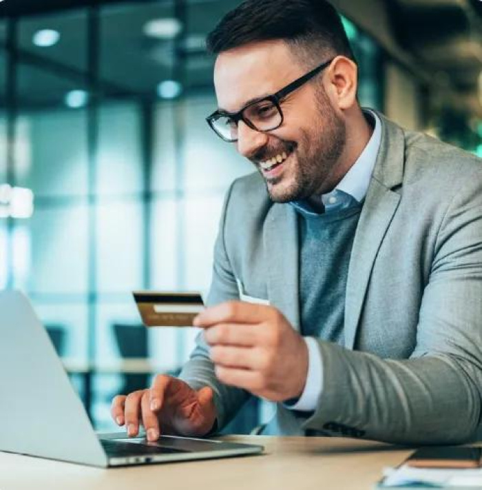 A small business owner in a suit smiling at his laptop while holding his business credit card from Essential Credit Union in Houston