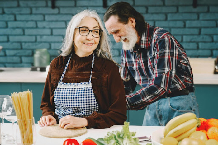 Elderly man helping elderly woman put on apron in kitchen before cooking